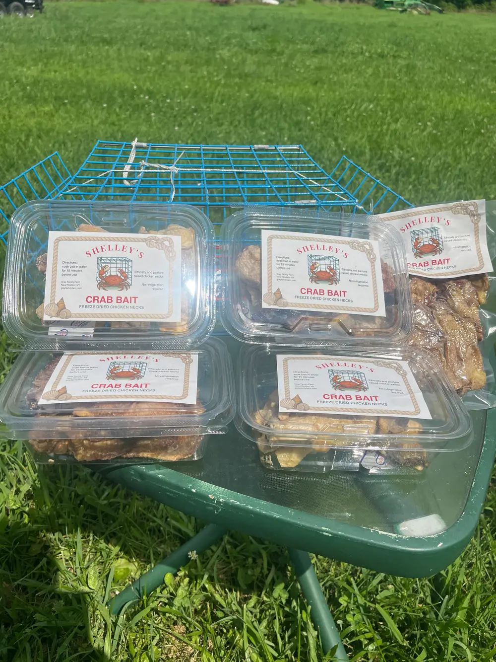 Containers of Shelley's Crab Bait on a wheelbarrow beside a blue crab trap, with pasture behind